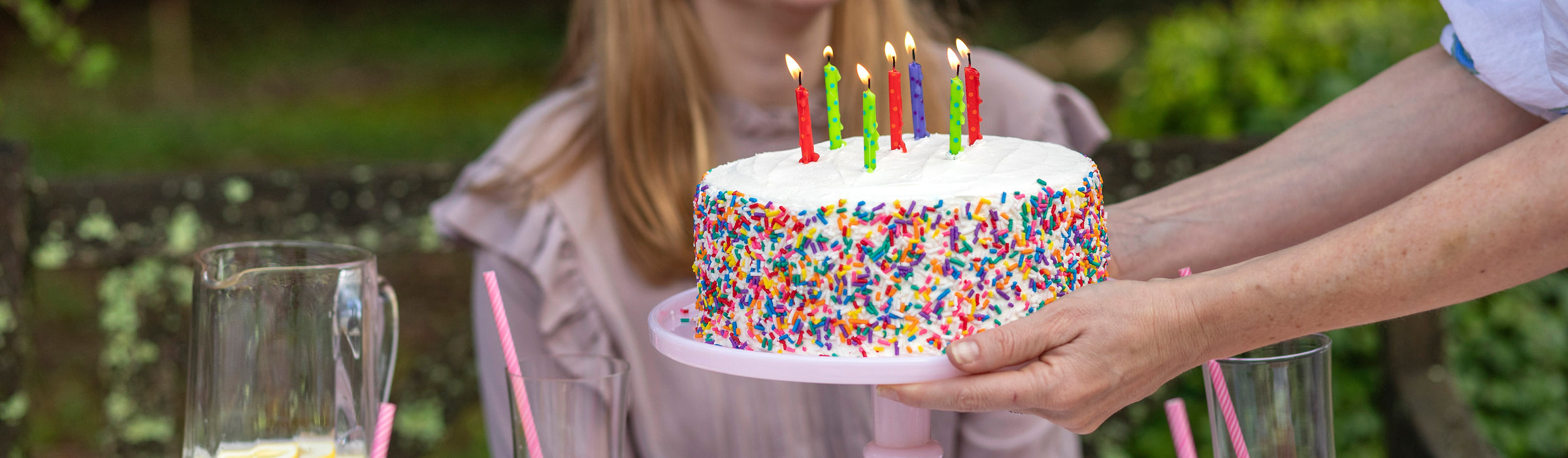Rainbow sprinkle birthday cake with lit candles carried to the table on a pink stand at an outdoor party