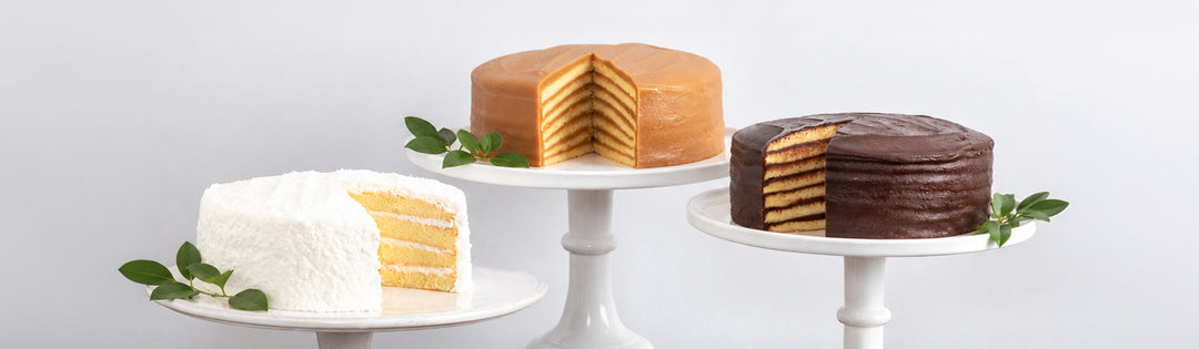 Coconut, seven-layer caramel and chocolate cakes on white pedestal stands, each cut to show the layers inside