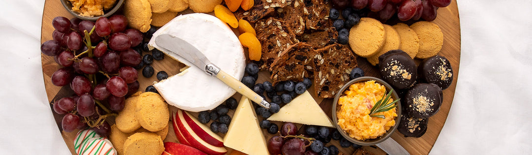 Overhead cheese board with brie, aged cheddar, pimento cheese, cheese wafers, grapes, blueberries, apple slices and chocolate-covered cake bites
