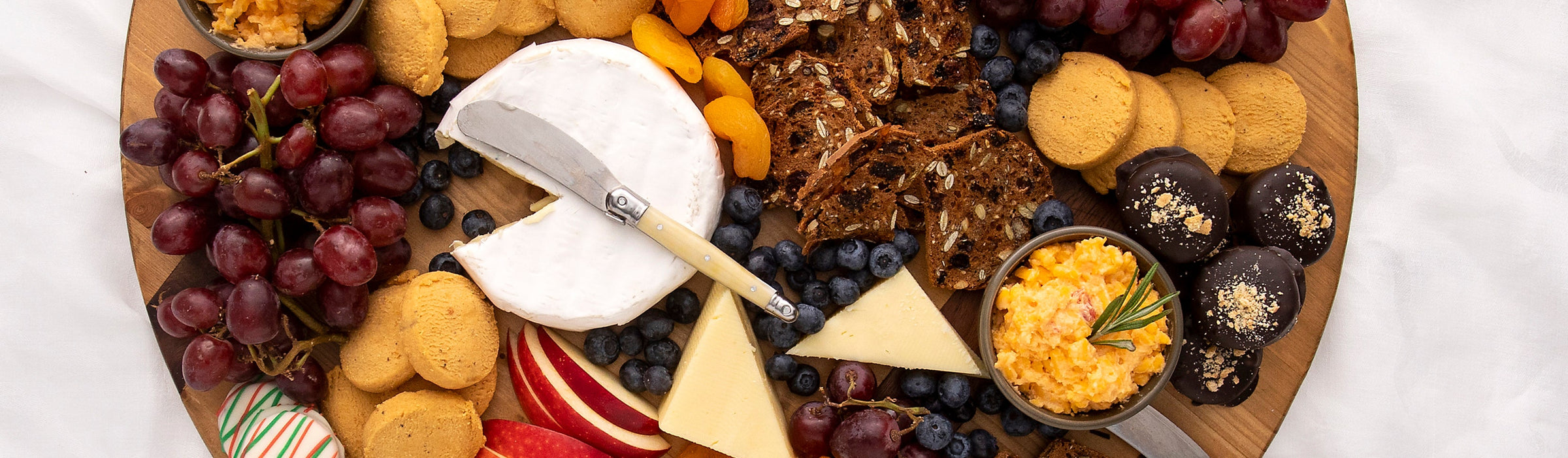 Overhead cheese board with brie, aged cheddar, pimento cheese, cheese wafers, grapes, blueberries, apple slices and chocolate-covered cake bites