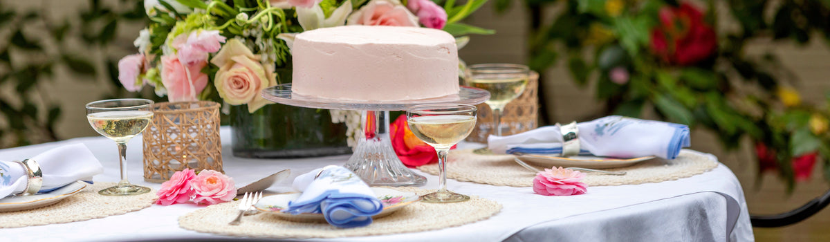 Pink-frosted cake on a glass pedestal at a garden table set with floral china, blue and white napkins and champagne coupes