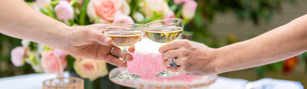 Two hands toasting champagne coupes over a pink sprinkle cake, with roses behind