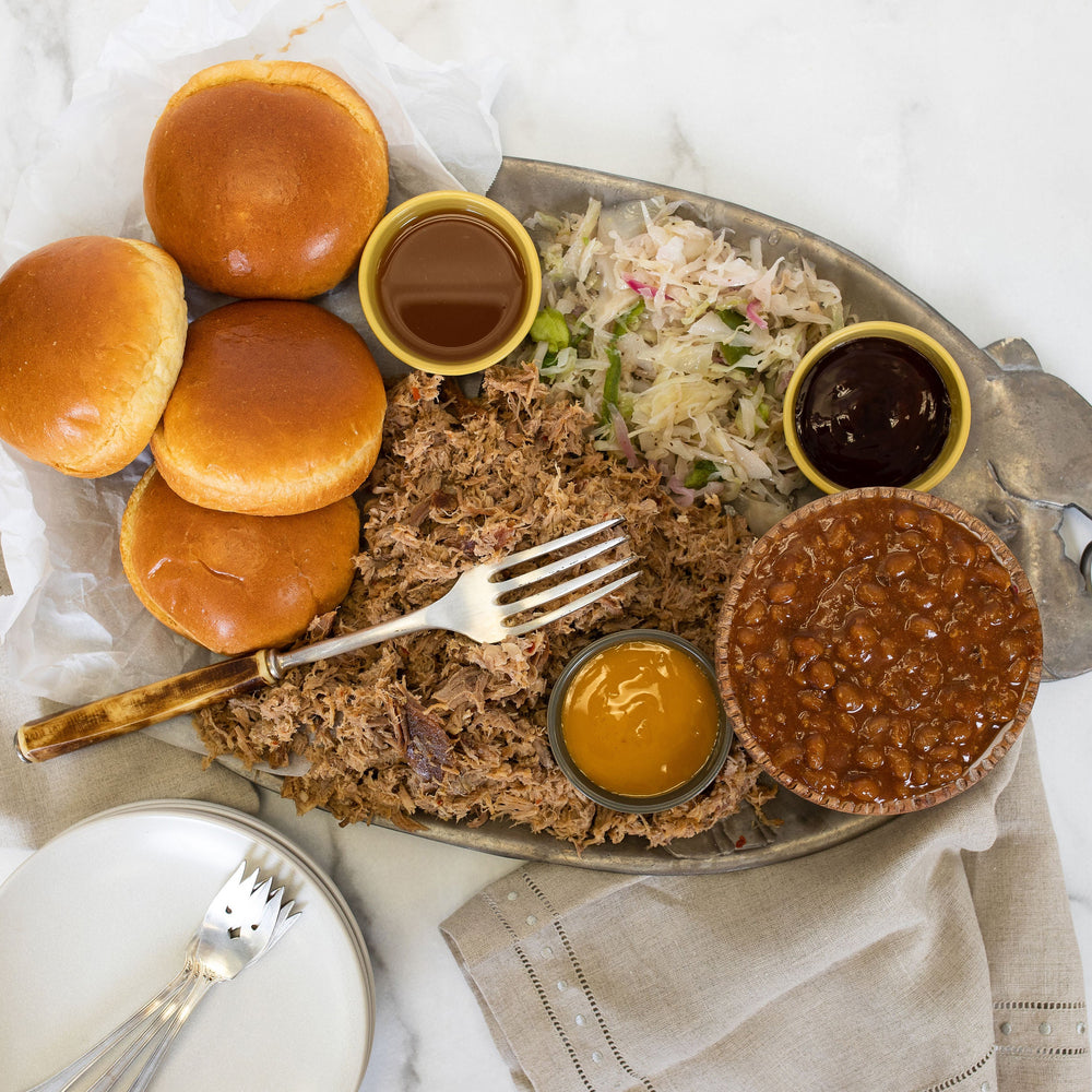 A pewter platter of pulled pork with buns, slaw, baked beans and three sauces.