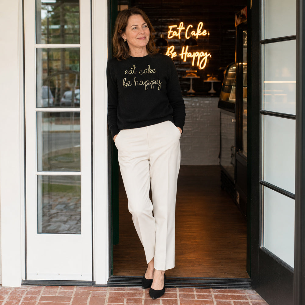 A woman in the black eat cake. be happy. sweater in the shop doorway, the neon Eat Cake. Be Happy. sign behind her.
