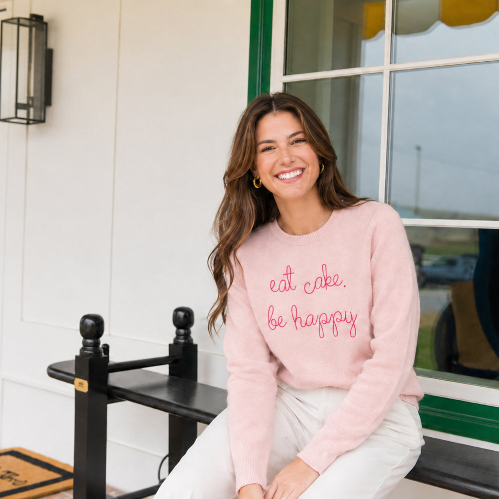 A woman seated on a black bench wearing the pink eat cake. be happy. sweater, on a white porch.