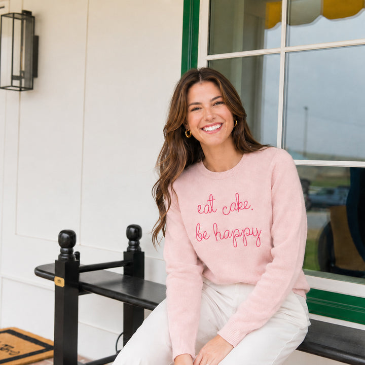 A woman seated on a black bench wearing the pink eat cake. be happy. sweater, on a white porch.